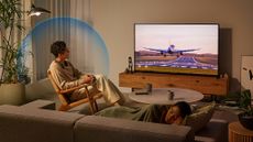 A man in a chair watching an aeroplane landong on TV. He's dressed in beige and is listening to a Sony Bravia Theatre U neck-worn speaker while another person sleeps on the sofa.