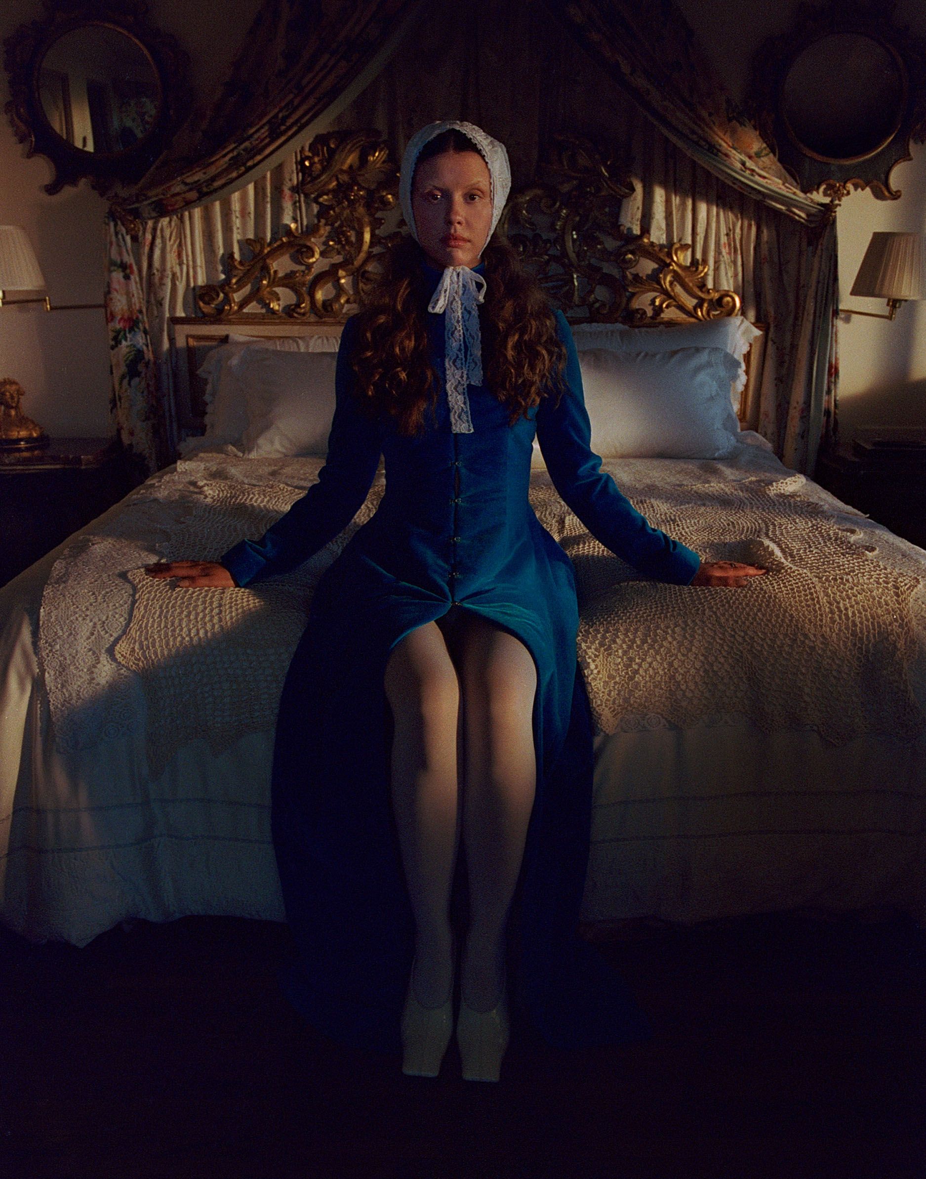 Mia Goth photo shoot for Who What Wear's October Cover Story. Mia Goth is sitting on a bed with ornate headboard. She is wearing a blue velvet long sleeve dress with white tights and a white lace headscarf.