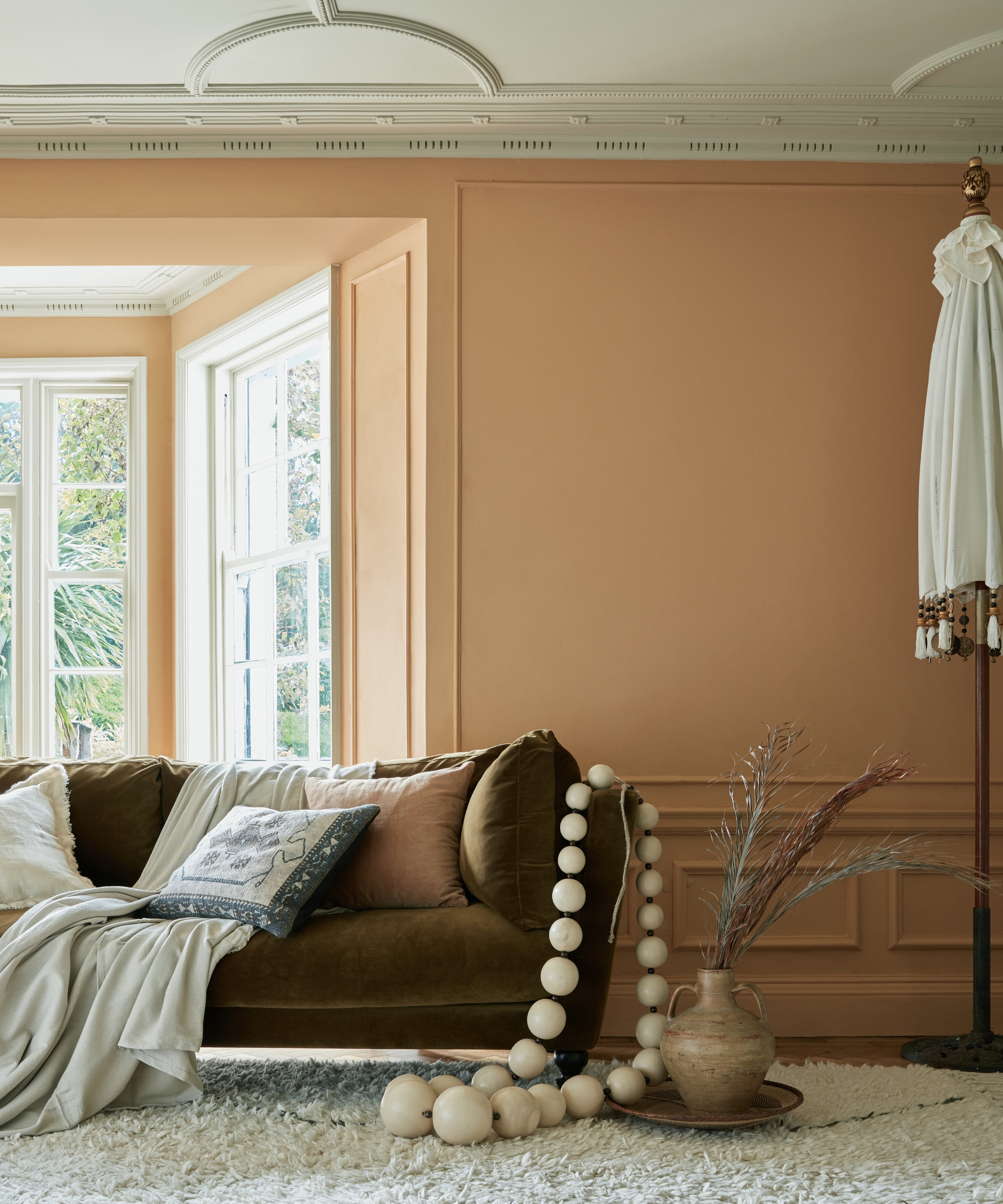 Warm terracotta wall with wood paneling, olive velvet sofa, wooden bead garland, and a fringed lamp.