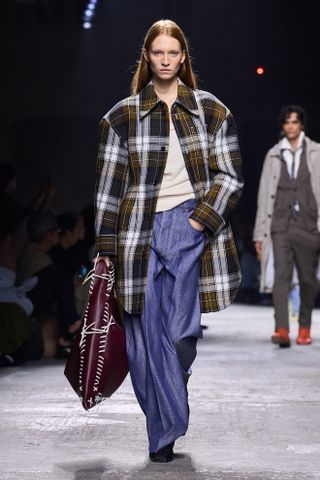 a model on Bottega Veneta spring 2025 runway wears a flannel outfit of a green, white, and blue plaid jacket, white top, and blue jeans