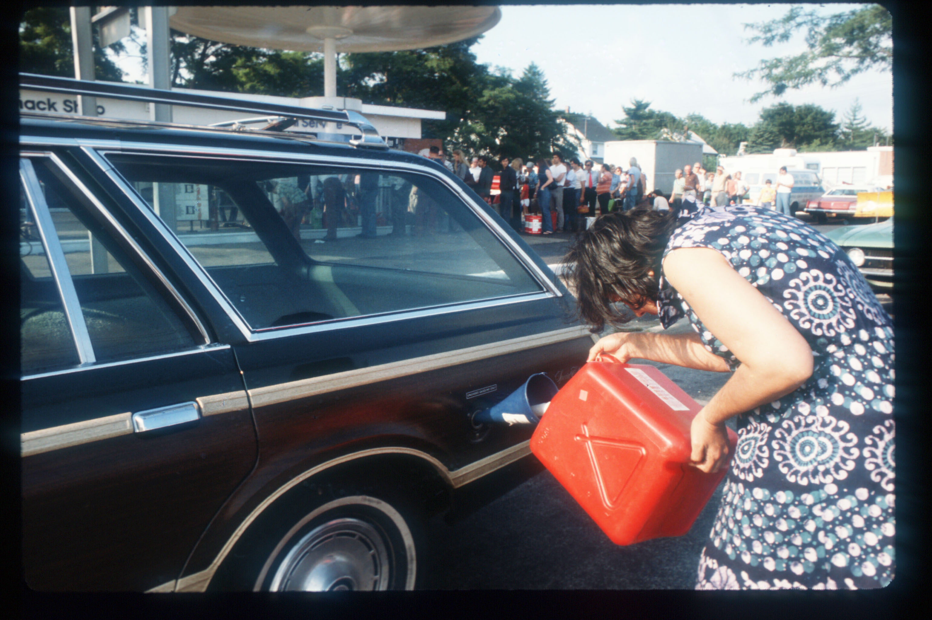 In the 1970s, a woman pours gas in the tank of her station wagon. A line of people waiting for gas stand in the background.