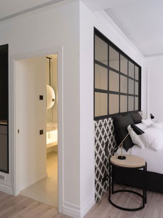 Interior view of a room at The Serras featuring wood flooring and white walls with a section behind the bed that has a black and white diamond pattern. The bed has black and white pillows and linen and there is a black grid style window above the bed. There is also a round wooden and black side table with a lamp and a partial view of another room