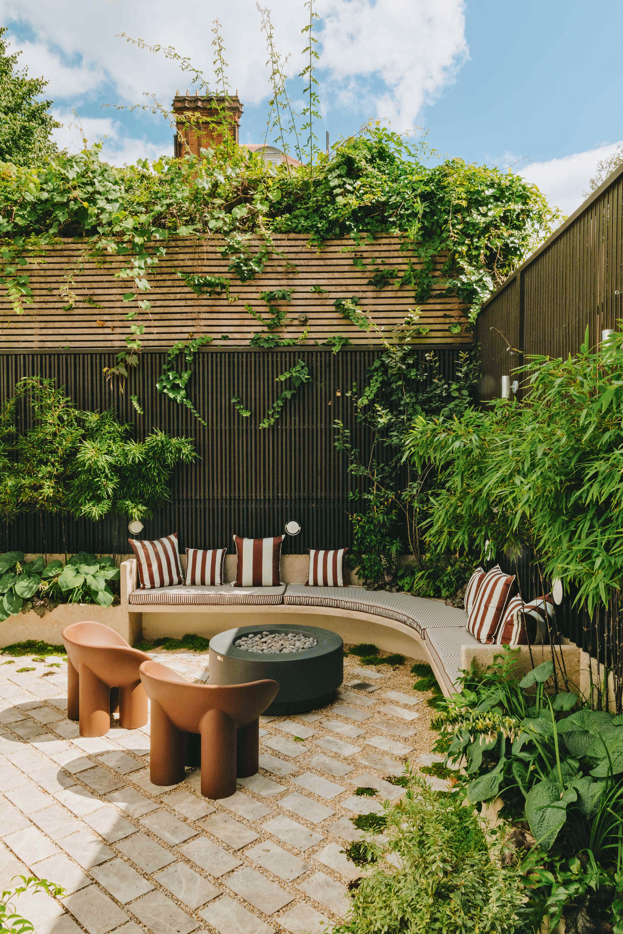 Corner of a garden with built-in curved stone bench, black firepit and terracotta Faye Toogood accent chairs