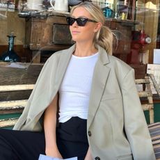style influencer Fredrika Ekerot sits on a wooden bench outside a shop in NYC wearing black sunglasses, an oversize tan blazer, white tank top, and black pants
