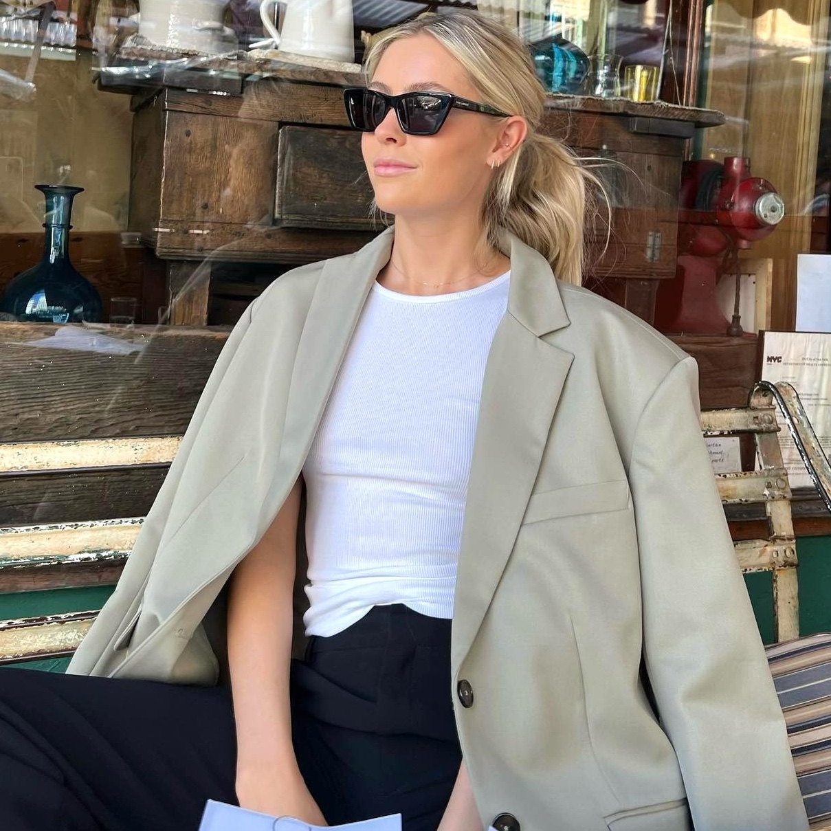 style influencer Fredrika Ekerot sits on a wooden bench outside a shop in NYC wearing black sunglasses, an oversize tan blazer, white tank top, and black pants