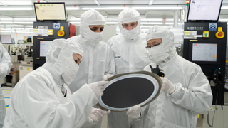 Four people in full white cleanroom suits and masks gather around to inspect a large, dark semiconductor wafer in a high-tech manufacturing facility