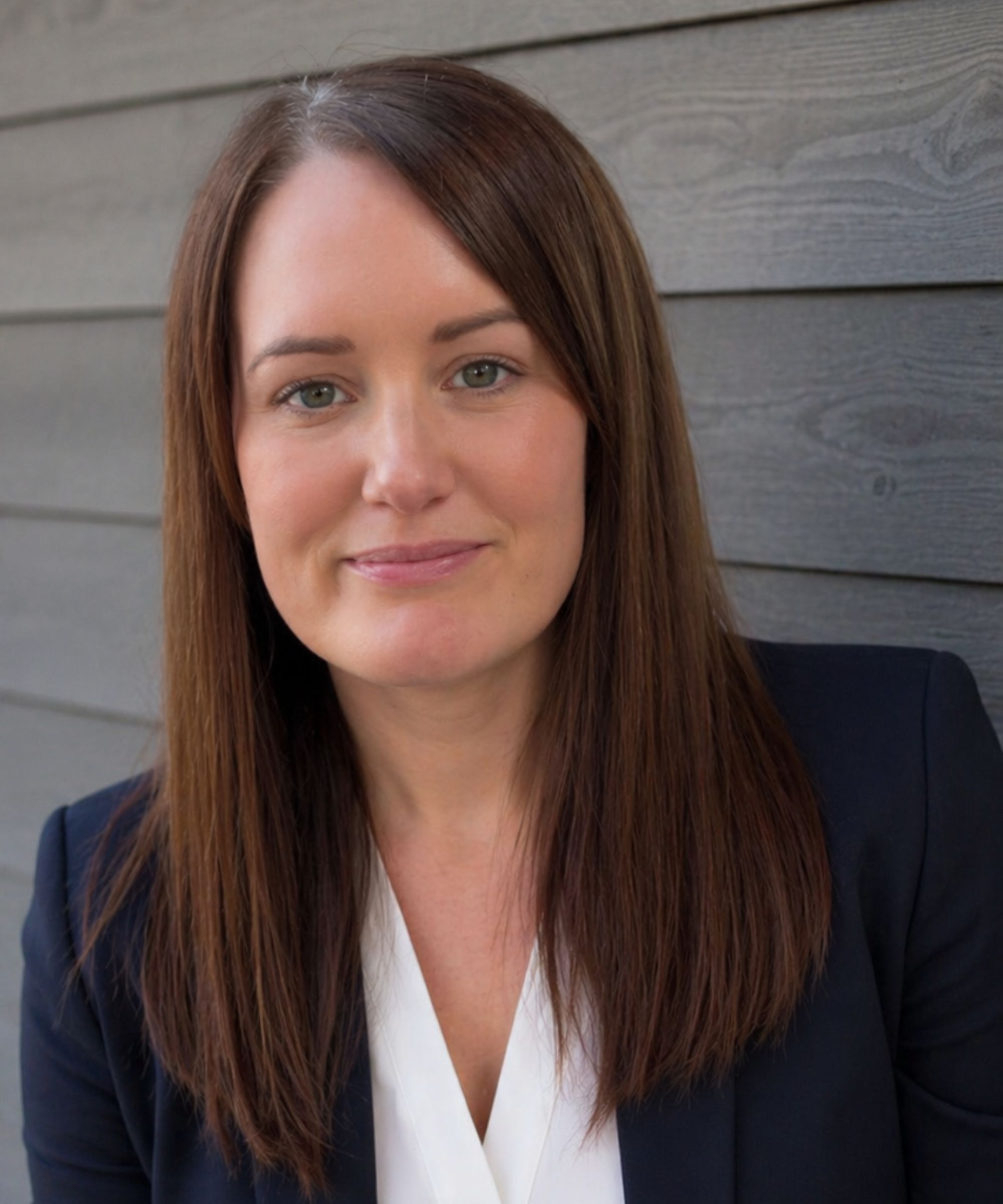Woman with long brown hair, navy blazer over white top, smiling softly at camera against grey wood-paneled wall.