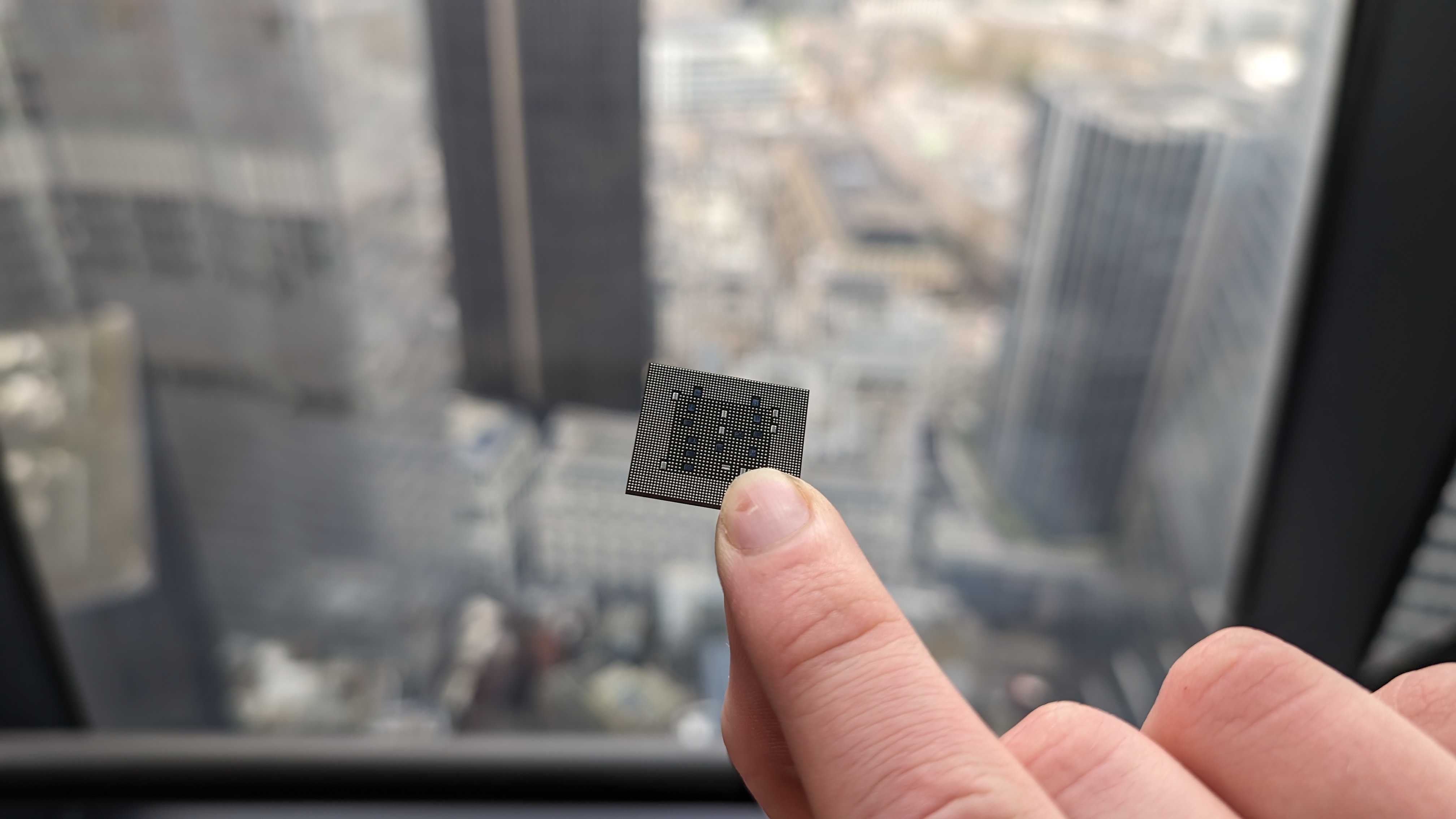 A Qualcomm Snapdragon X chip being held carefully in two fingers against a cityscape background.