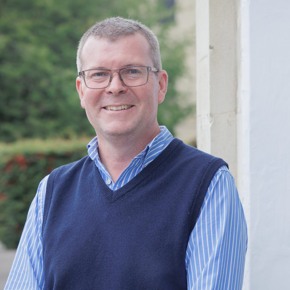 male with short hair, glasses wearing blue shirt and jumper stood outside