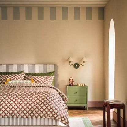 Habitat Wiggle Metal 2 Light Wall Light - Green & Natural hanging on beige wall above green bedside table, beside green striped headboard with red patterned sheets