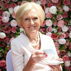 Mary Berry sitting drinking a cup of tea in front of a large backdrop of roses