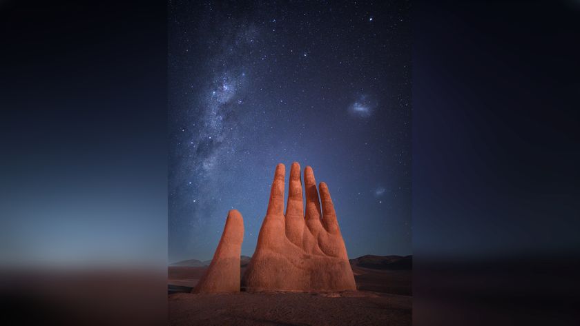 A large, surreal sculpture of a hand rises from the desert, under a starry night sky illuminated by the Milky Way