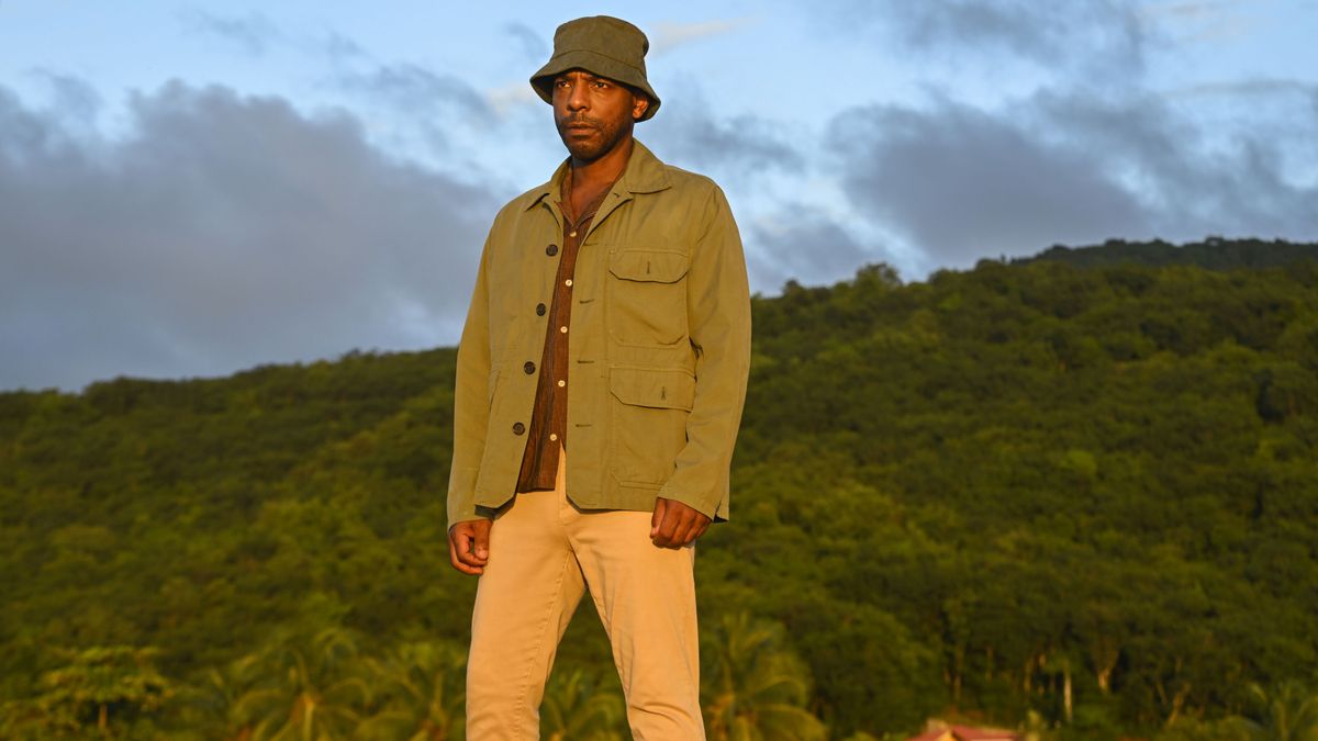 DI Mervin Wilson (Don Gilet) stands on the beach in the late evening light, the landscape behind him a canopy of trees with grey clouds in the sky. He is looking out to sea, wearing a light canvas jacket and a green bucket hat over his clothes, and looking crestfallen.