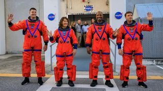 four astronauts in orange spacesuits waving at the camera in front of a doorway festooned with nasa logos