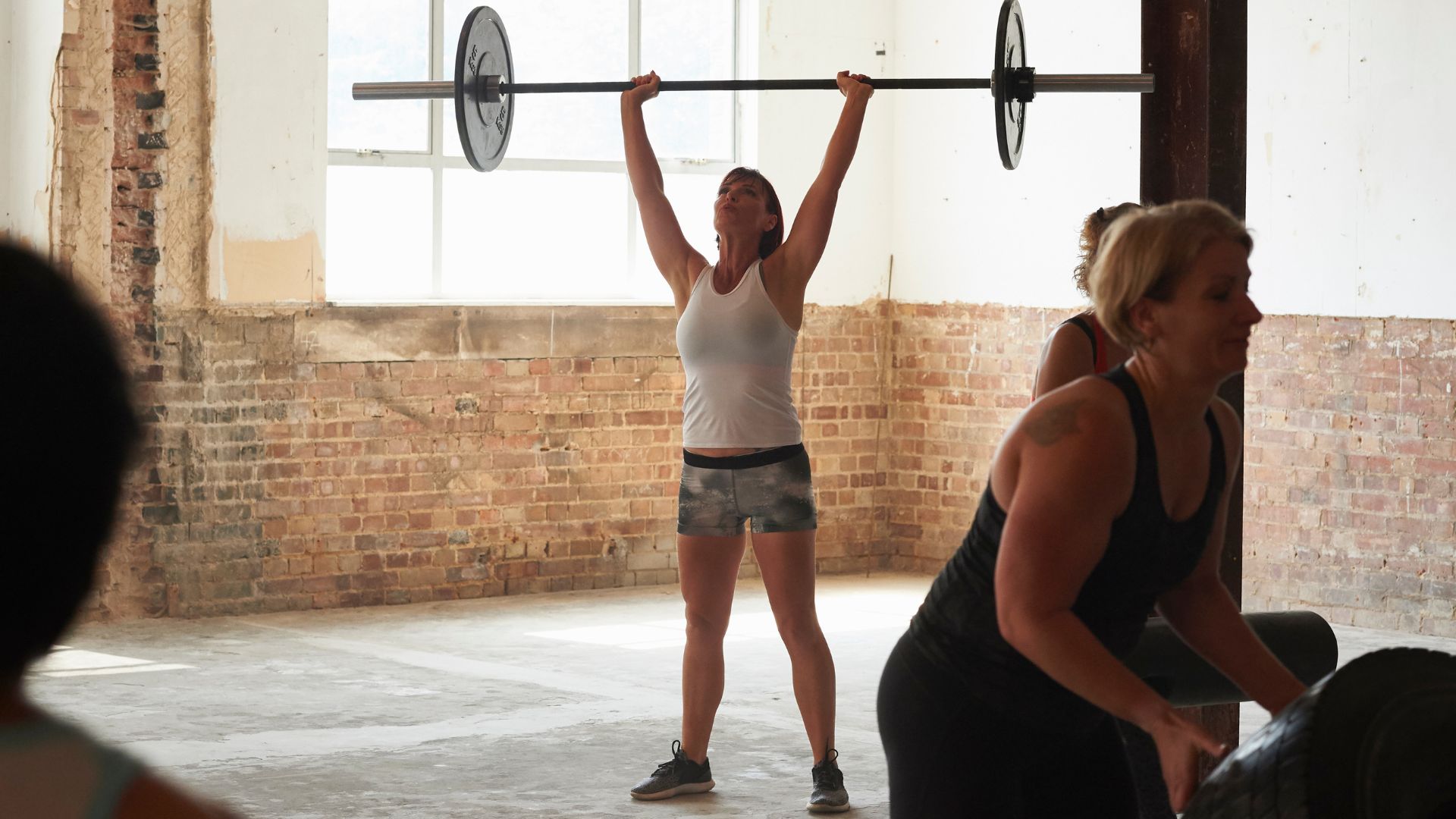 Woman lifting barbell above head with weight on either side wearing gym clothes and standing with friends in foreground