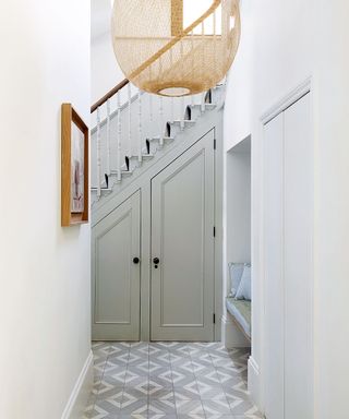Hallway with the staircase and built in cupboards, large woven lampshade and grey and white tiled floor.