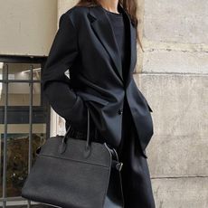 Woman wearing black blazer with black t-shirt, black slacks, and The Row Margaux bag, standing on Paris street.