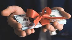 A pair of hands hold a bundle of money tied with a red ribbon.