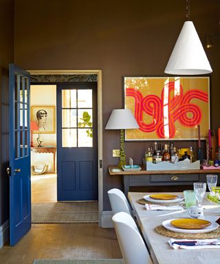 dining room with mid brown walls, blue doors, 1960s fluorescent statement artwork, gray and wood console, white washed wood table and white modern chairs