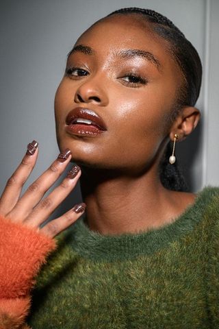 a model backstage at Ahluwalia spring/summer 2025 show with her hand raised wearing brown 3d nail polish, gold pearl earring and green and orange sweater