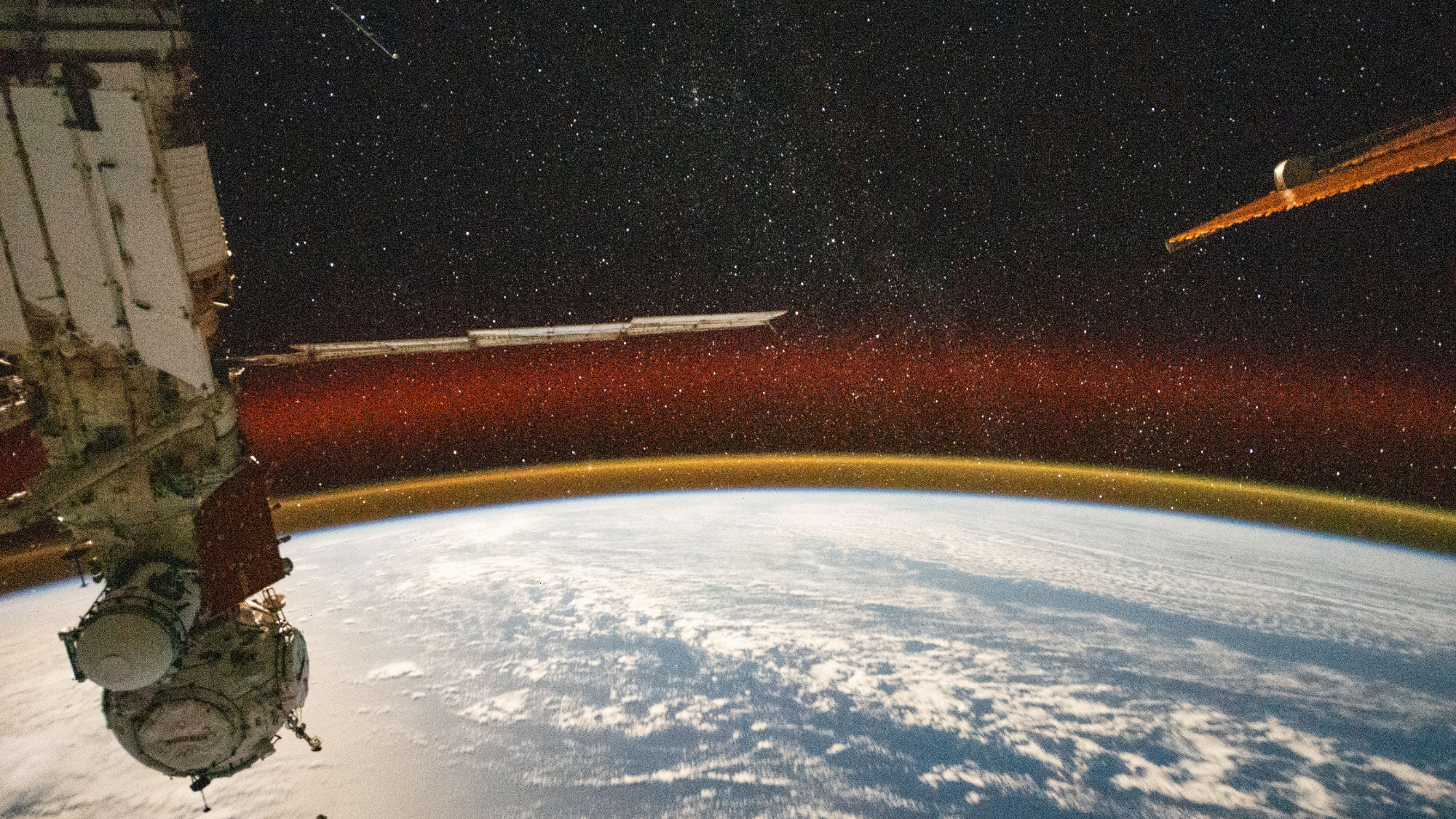 the round edge of earth is seen blue with a swath of clouds, beneath a thin orange layer of atmosphere crowned in a red glow with the black starry sky of space above. in the foreground on the left, a russian space station capsule hangs downward. On the right, the golden orange reflective edge ot a solar panel slices the stars.