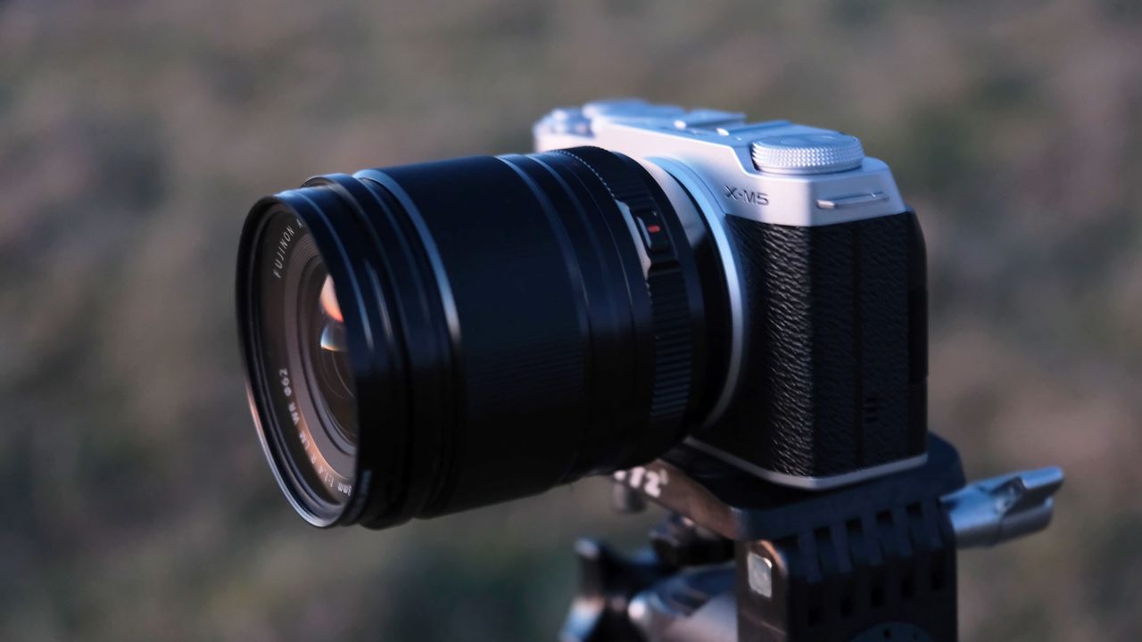 A side view of the Fujifilm X-M5 with the Fujifilm XF 18mm f/1.4 lens attached and a field in the background