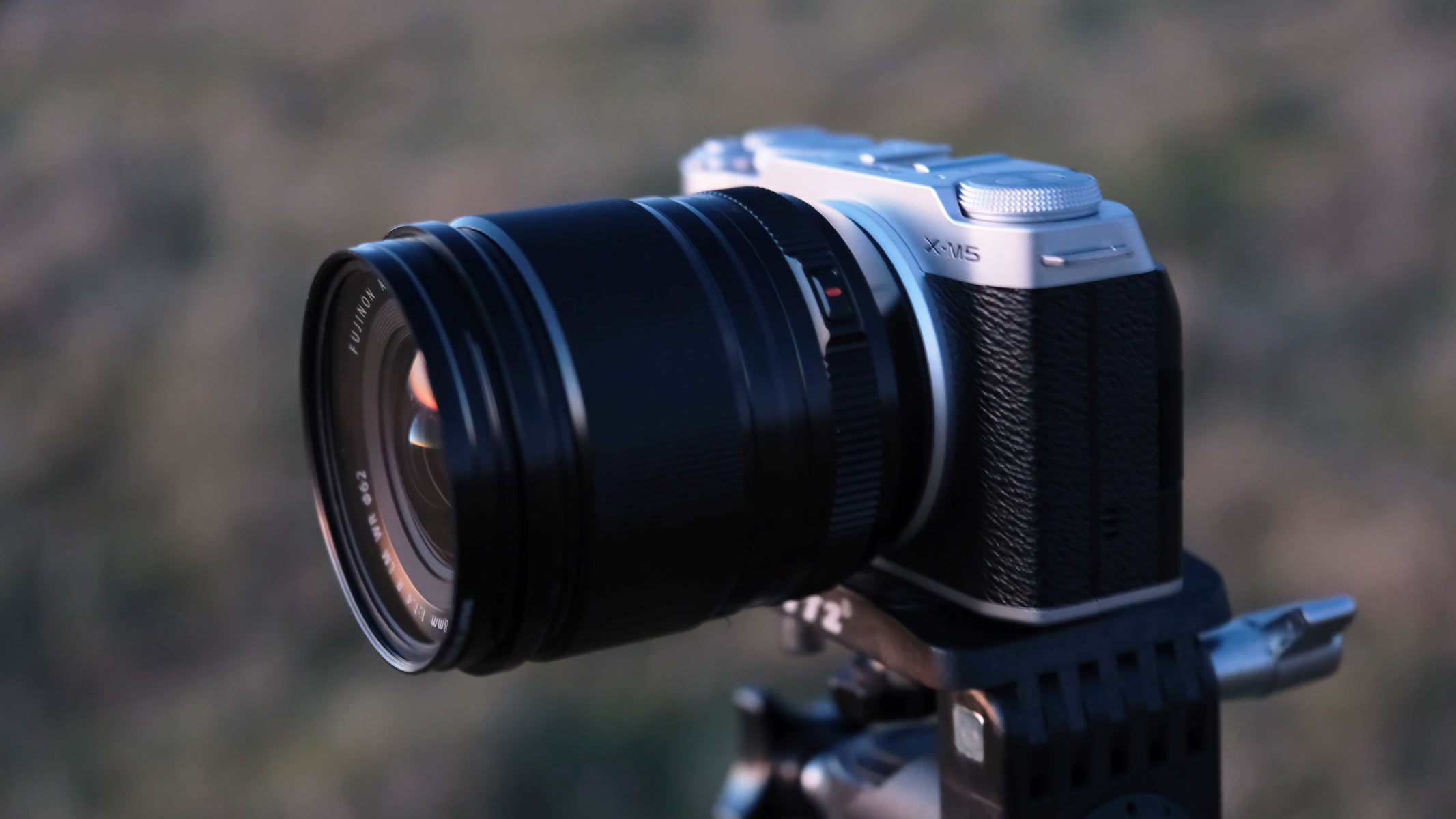 A side view of the Fujifilm X-M5 with the Fujifilm XF 18mm f/1.4 lens attached and a field in the background