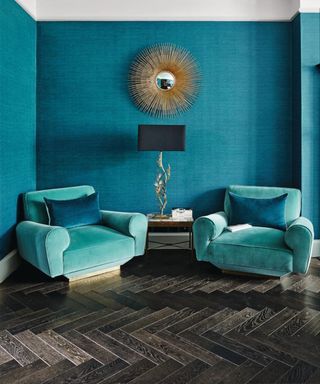 Two bright aqua armchairs on a dark wooden floor with bright blue walls, gold circular mirror and white cornices.