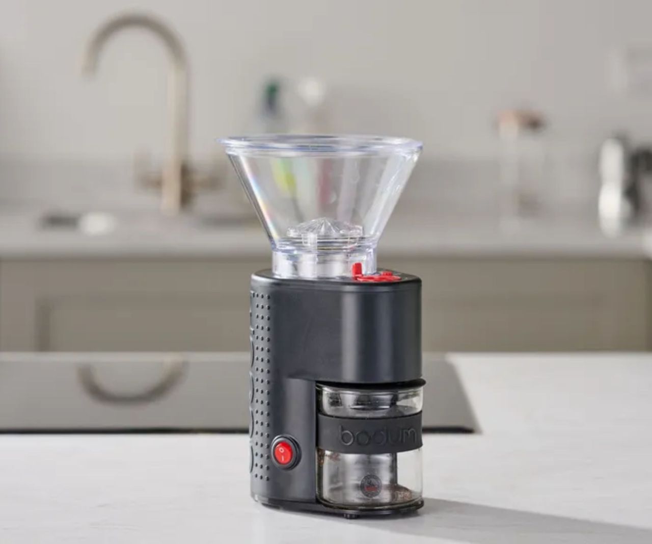 A compact black coffee grinder with a clear plastic funnel on the top, an out of focus kitchen in the background.