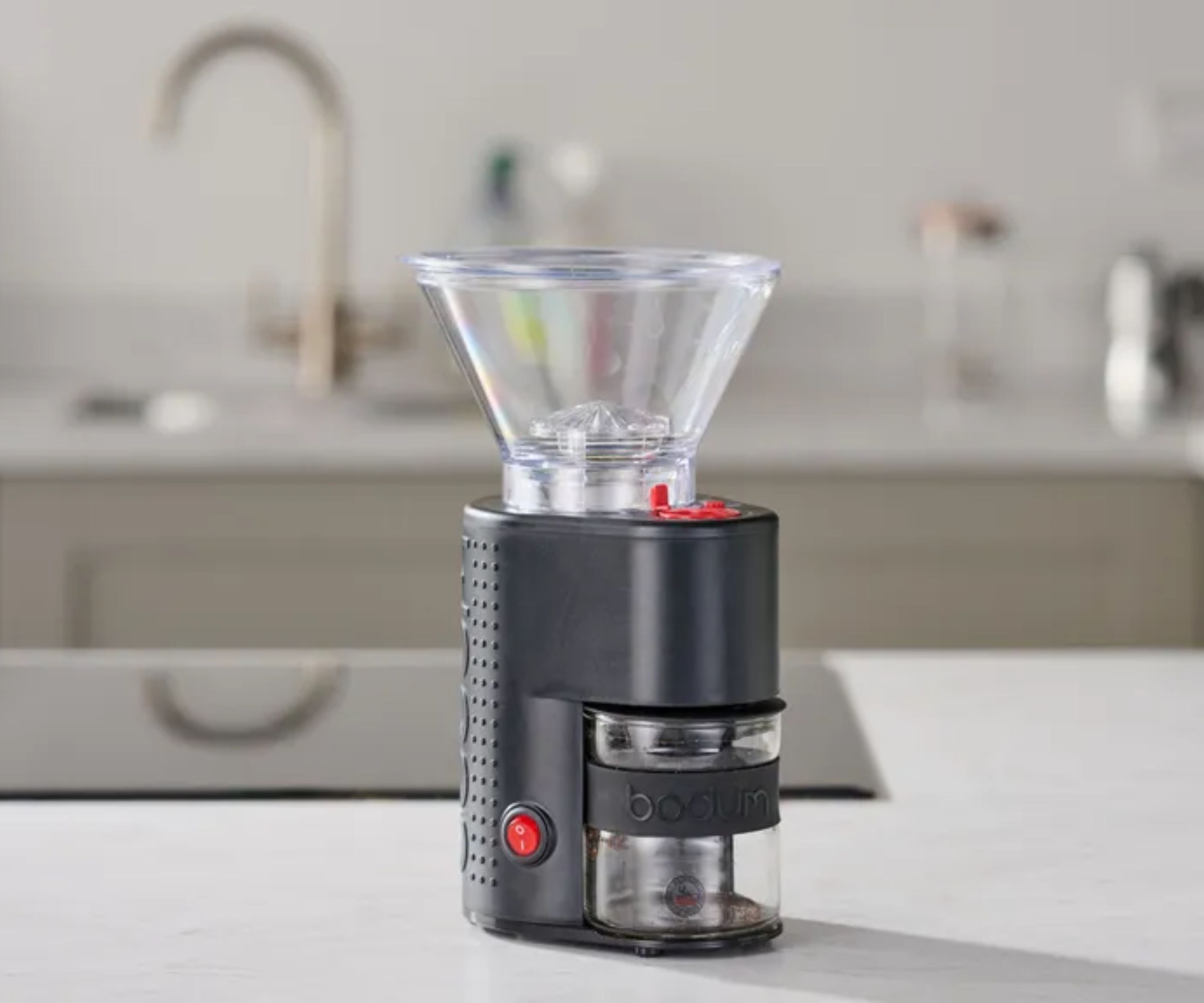 A compact black coffee grinder with a clear plastic funnel on the top, an out of focus kitchen in the background. 