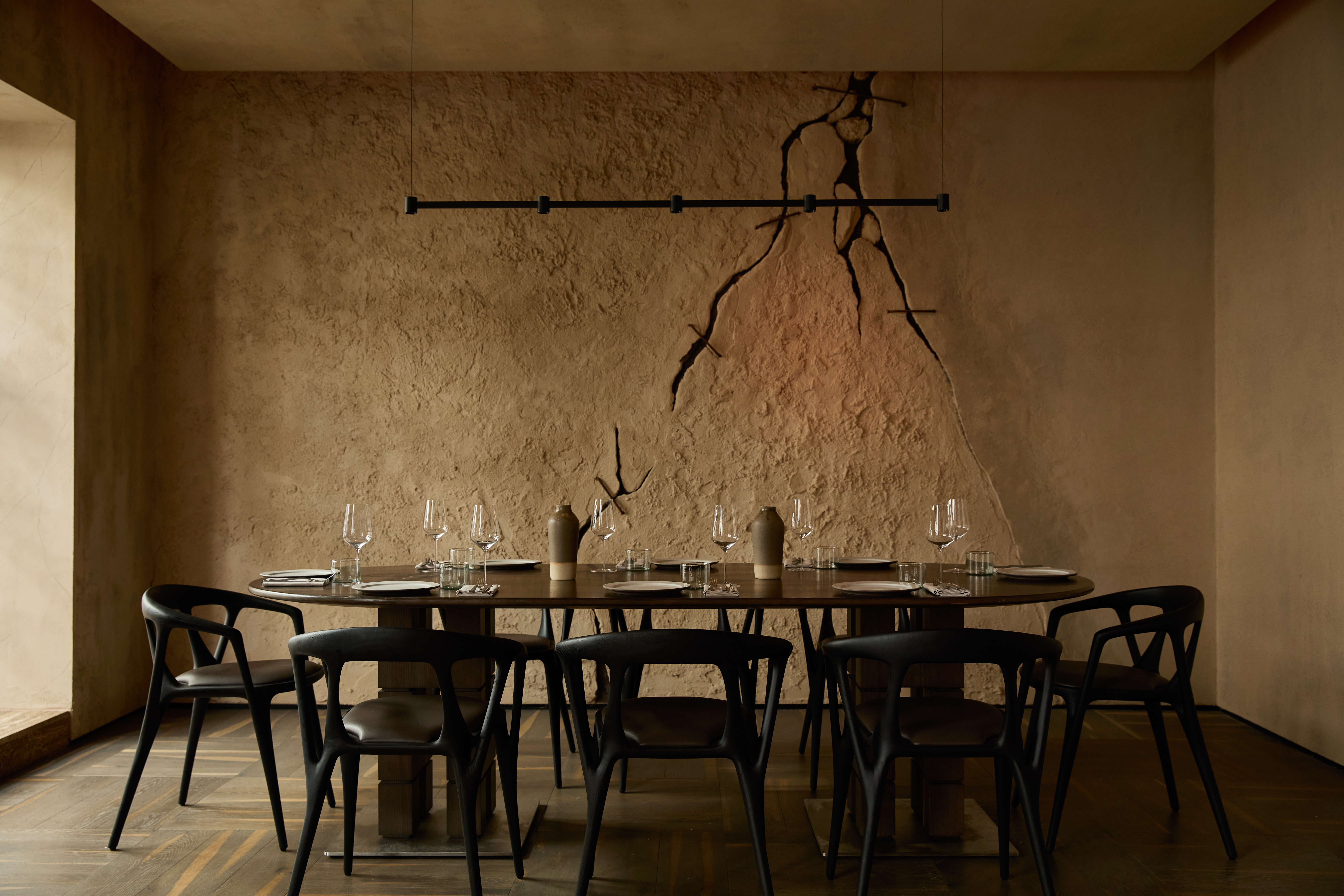 A private dining room in a brutalist restaurant featured cracked cement walls, a minimalist pendant light in wrought iron, and wooden furniture as well as floors.