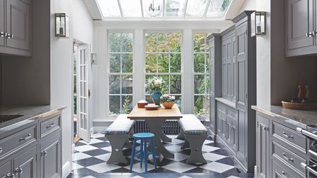 A black and white tiled kitchen with dark grey cabinets, a wooden dining table with grey benches, and large white windows.