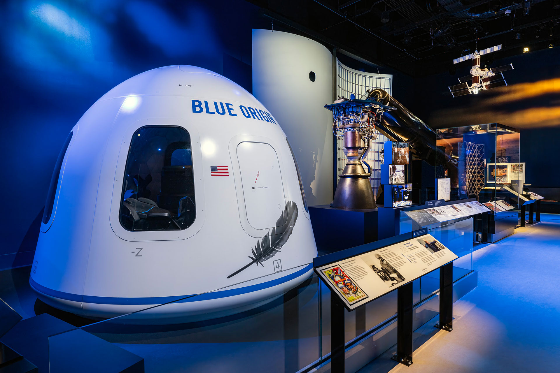 a space capsule sits in the foreground of a museum gallery with other rocket parts on display