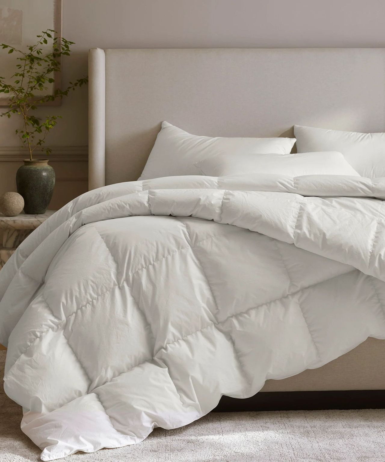 A white Boll &amp; Branch Down Duvet Insert pictured with white pillows on a cream upholstered bed, in a neutral bedroom. The walls are a pale pinky grey, and to the left og the bed is a marble bedside table with a large green vase with a plant, and a small circular stone ornament. Above the table, the corner of a wooden frame with a white artwork is seen, and in the foreground is a pale beige neutral carpet.