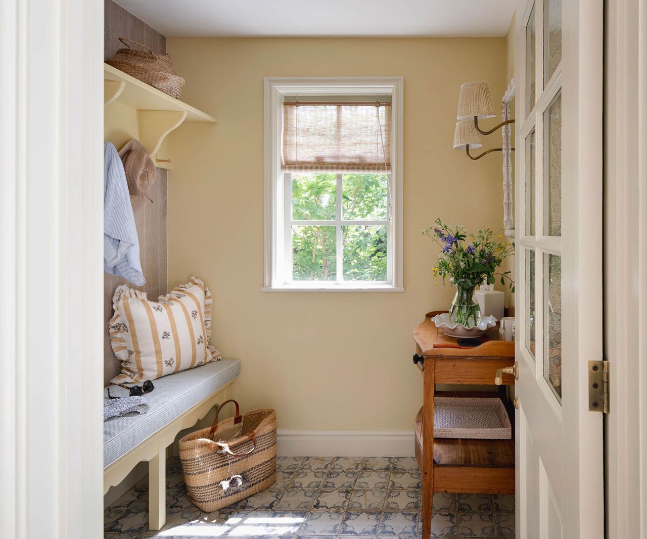 Yellow boot room with mudroom bench, cushions, wicker basket, patterned tiled floor, window with green view
