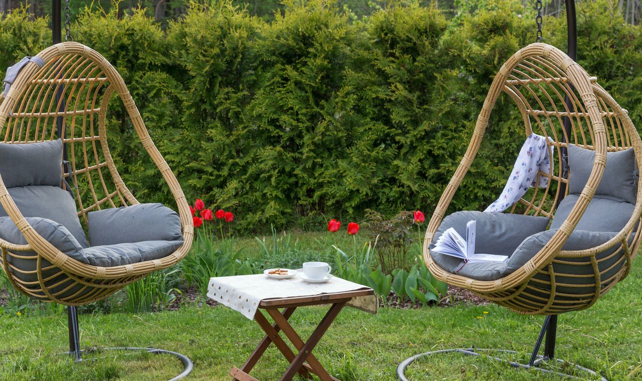 Two rattan egg chairs in between a bistro table in a green backyard with red flowers