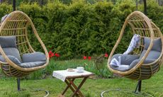 Two rattan egg chairs in between a bistro table in a green backyard with red flowers