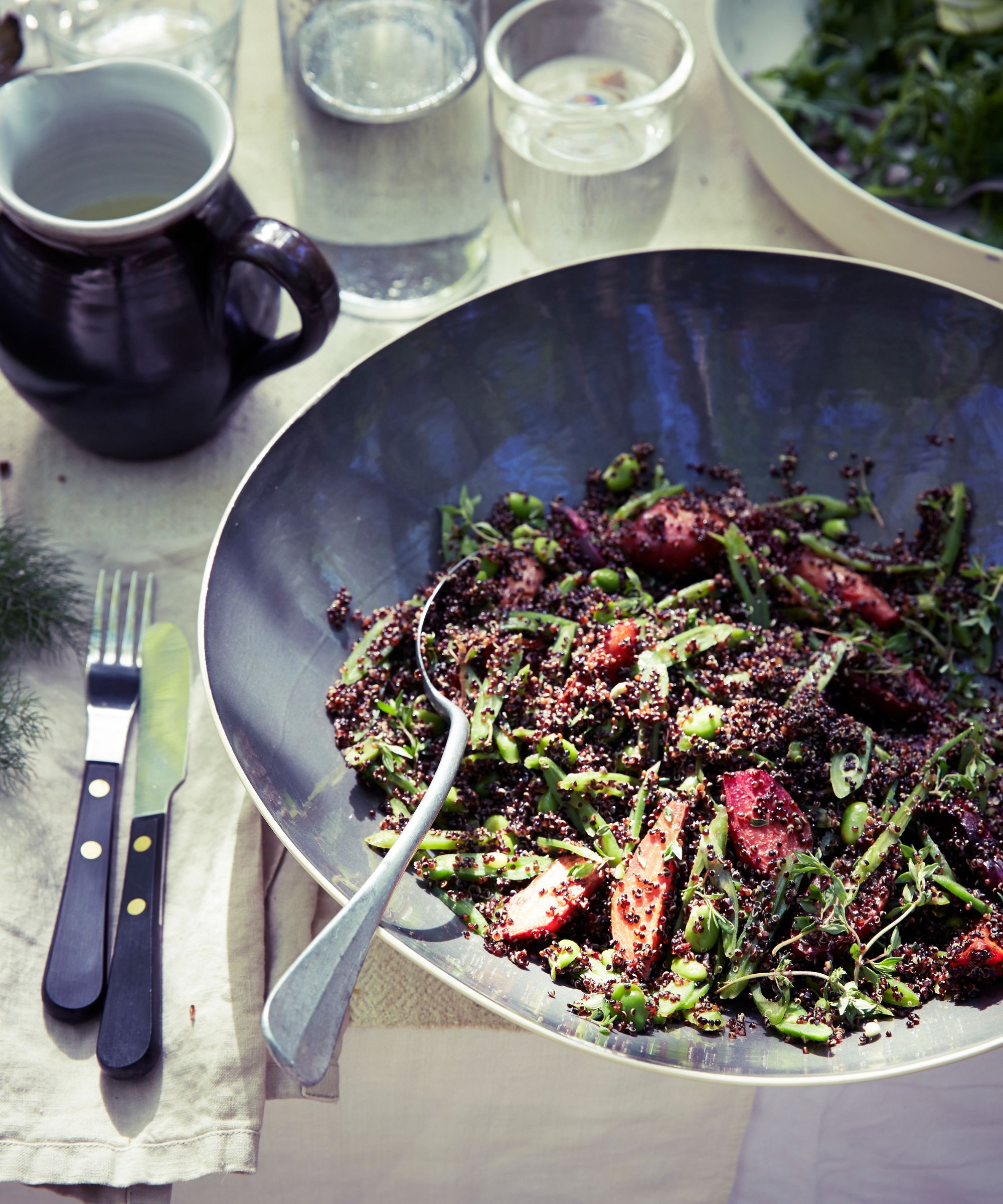 Black quinoa and beet salad in a large grey bowl on a table with knife. fork, jug, water glass and linen napkin