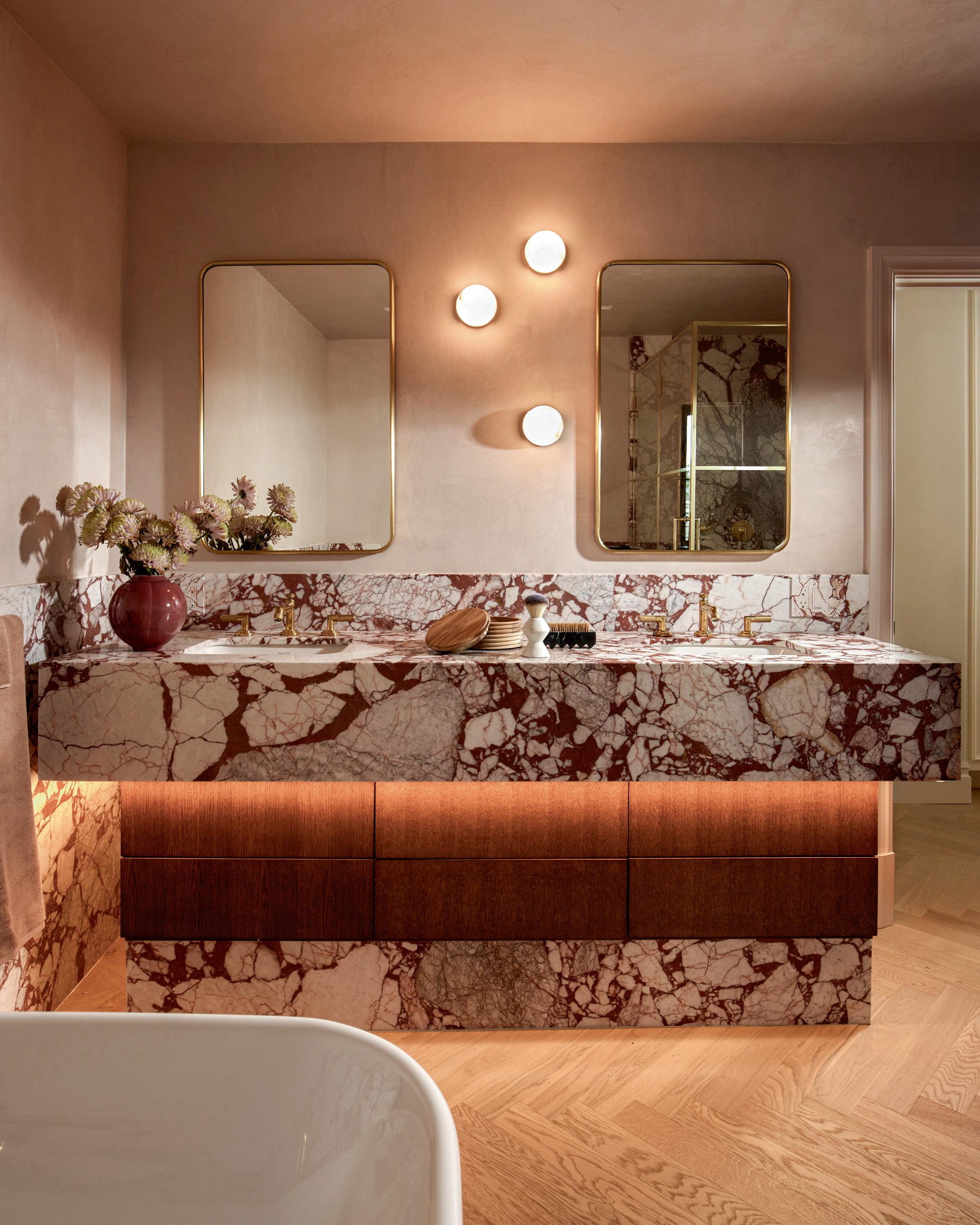 A bathroom with dusty rose Roman clay walls, Calacatta Viola Rosso marble vanity and panels, gold framed mirrors and simple wall sconces