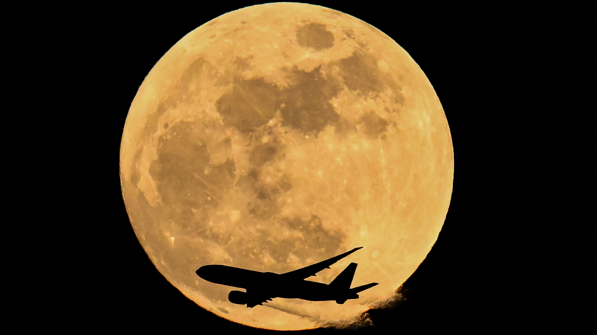 A yellow full moon glows in a black sky as the silhouette of a passenger jet crosses its lower half.