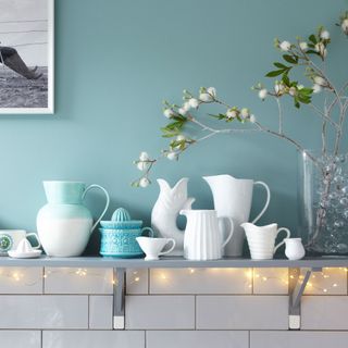 Kitchen makeover with white metro tiles blue wall, fairy lights, vase of flowers and a collection of white crockery