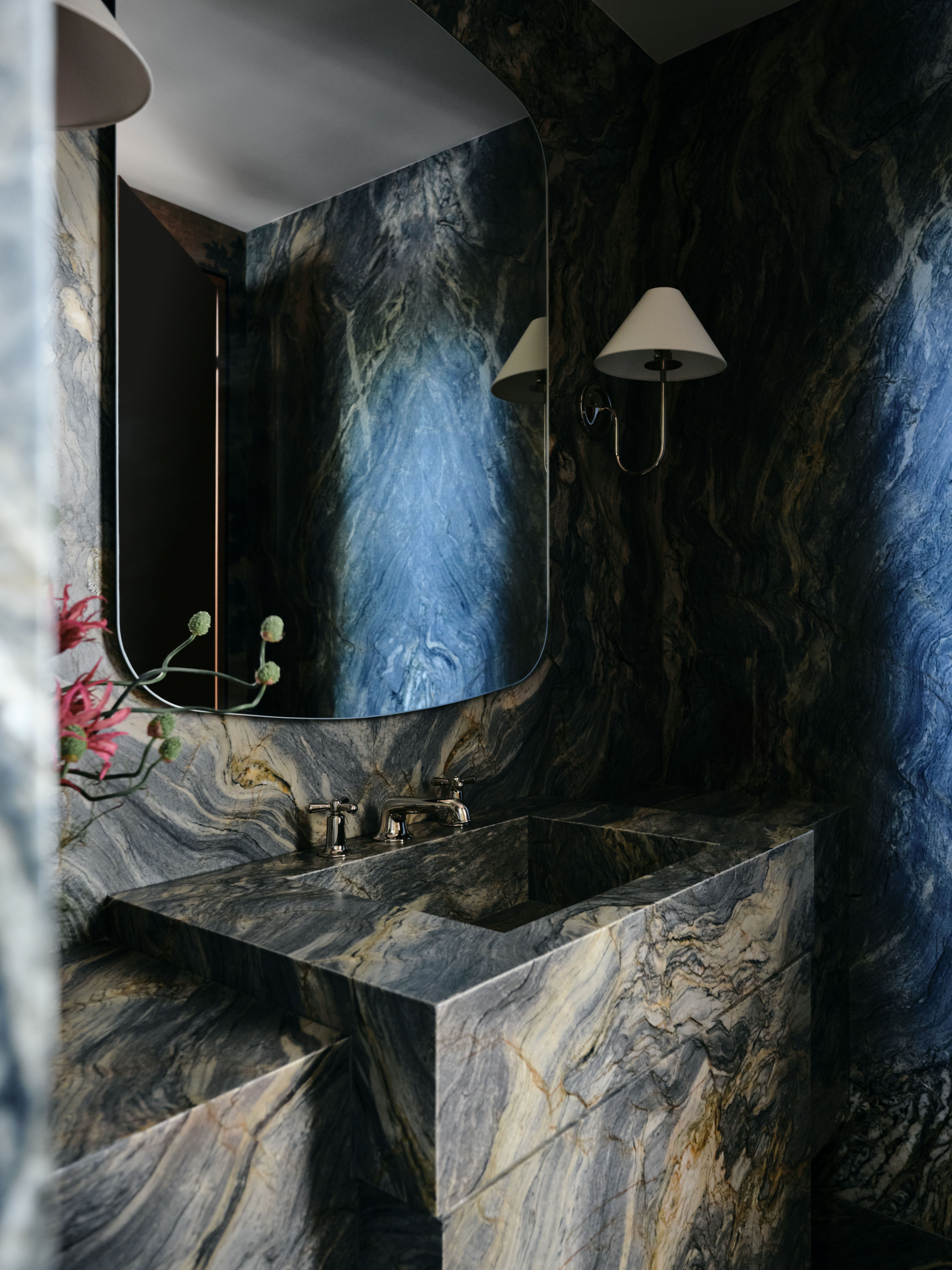 Black marble-clad bathroom, seen diagonally and decorated with traditional white sconces with brass arms, flowers, and brass fixtures.
