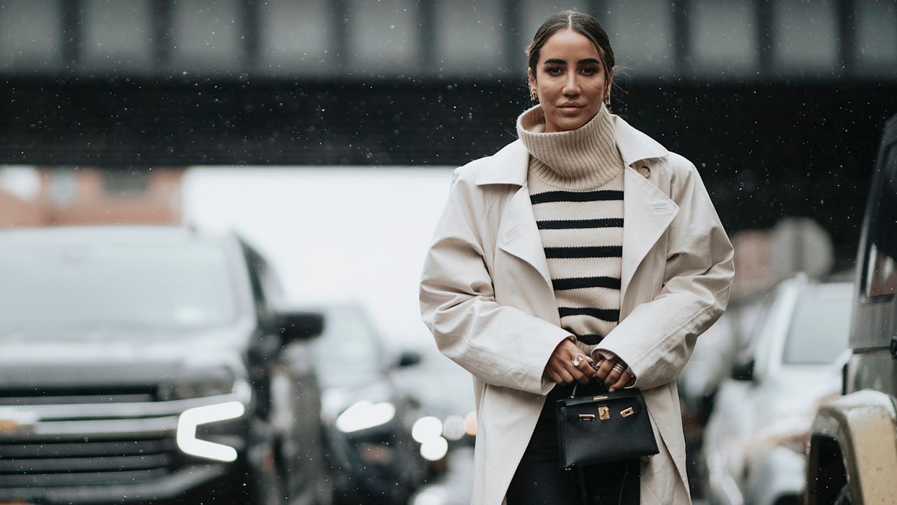 woman wears striped beige sweater with trench coat and black handbag 