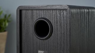 a pair of black wood speakers with a yellow kevlar cone photographed on a wood desk with a computer and blue wall and a plant in shot