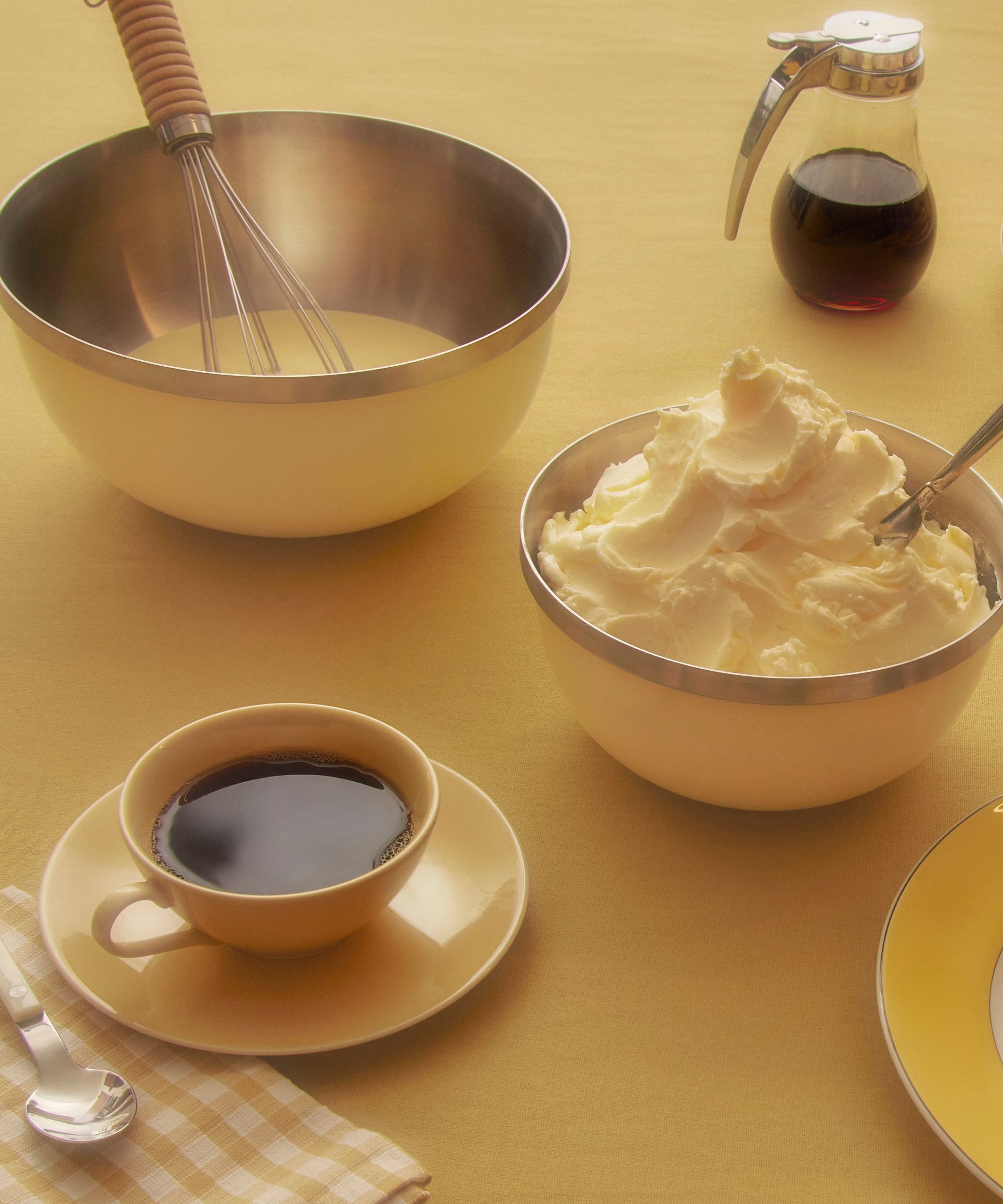 Butter yellow stainless steel mixing bowls on a yellow table beside a yellow coffee mug and saucer.