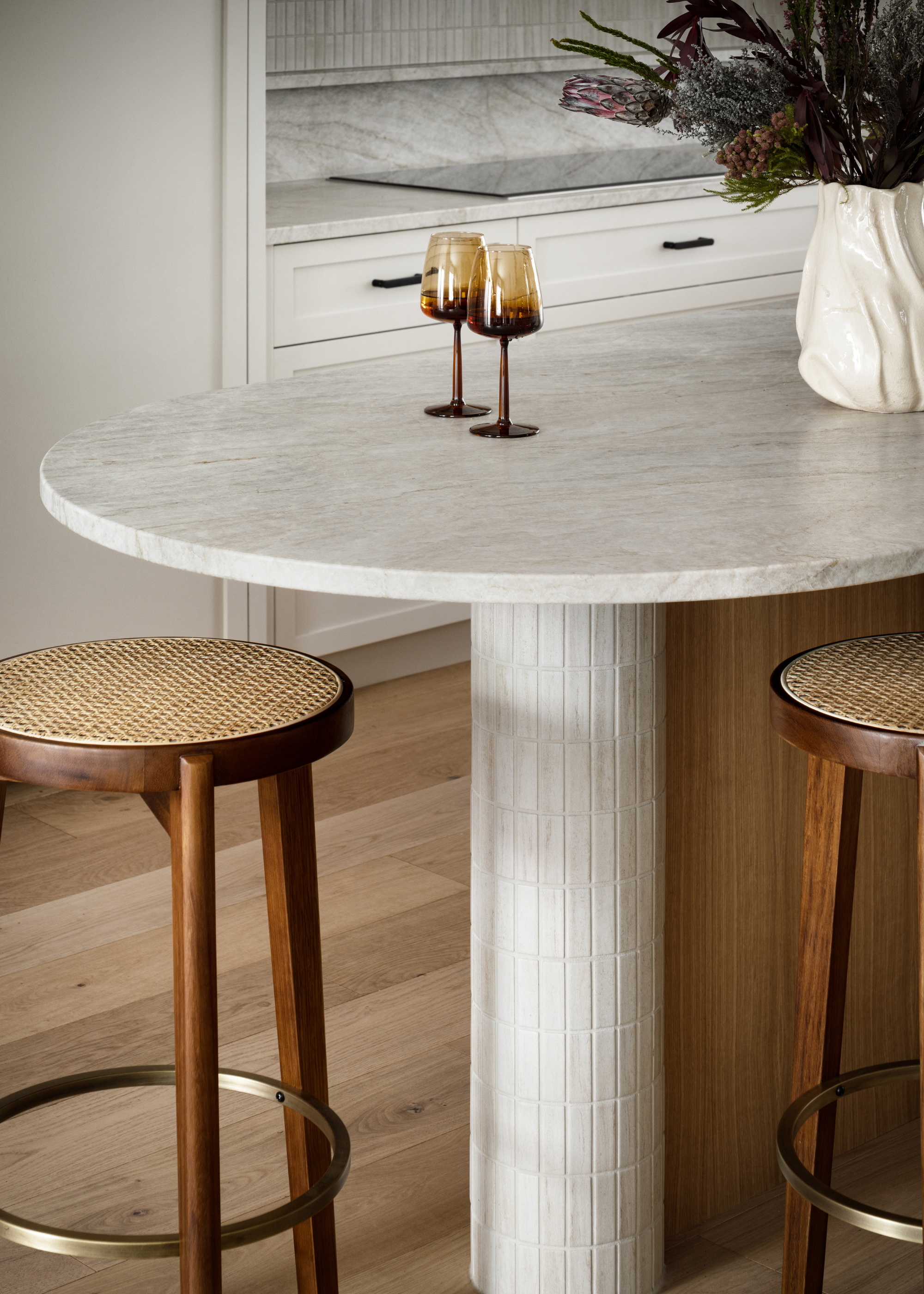 A close-up of a stone kitchen island with a pair of amber wine glasses near a vase of flowers alongside wood and rattan stools