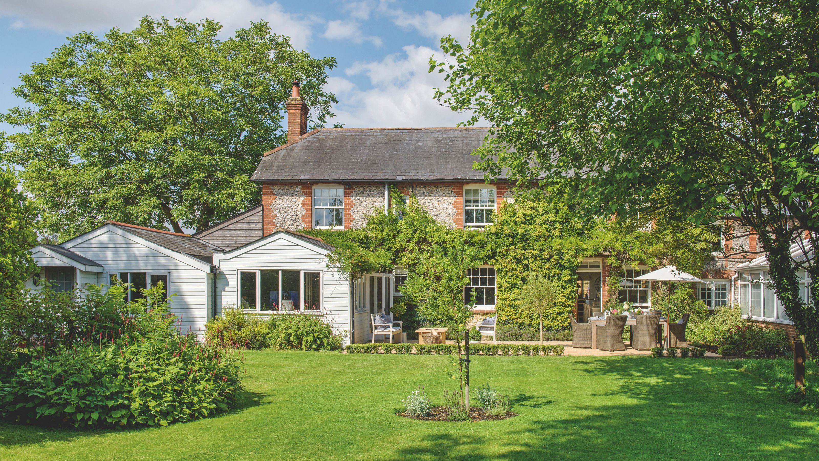 Large garden laid to lawn behind a large cottage-style house, with lots of trees in the garden