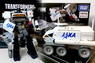 closeup of a six-wheeled white moon rover transformer toy with the words "jaxa" and "toyota" written on its side, in front of the box it's sold in.