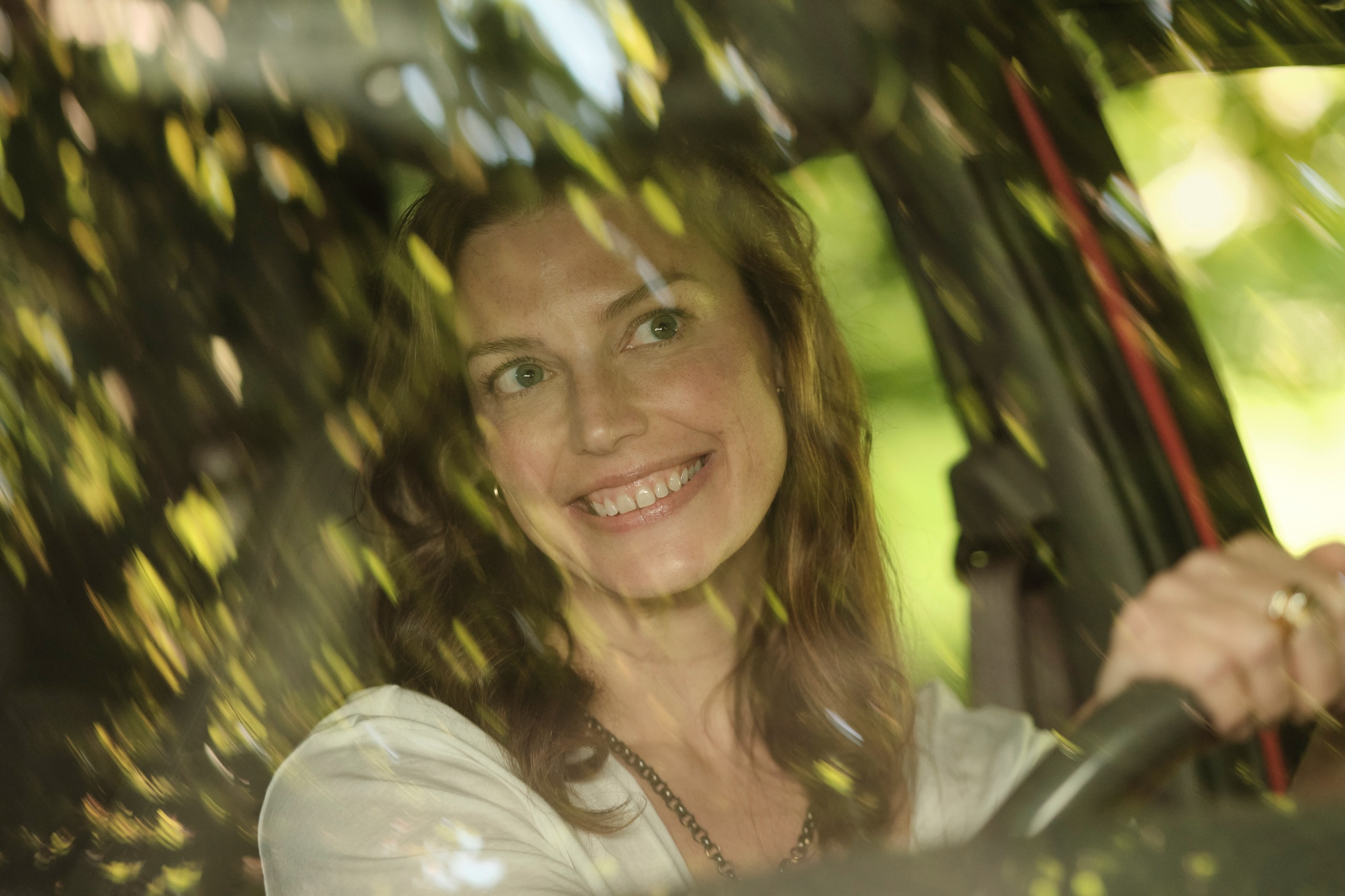 A smiling woman (Jessica Par&amp;eacute; as Ella Tao) is seen in a car's driver seat through a windshield obscured by the reflection of a tree, in Netflix's 'Crew Girl.'