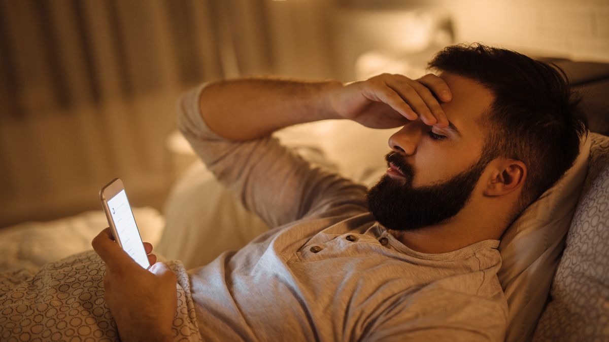 A young man is laying in bed looking at his phone, he looks stressed